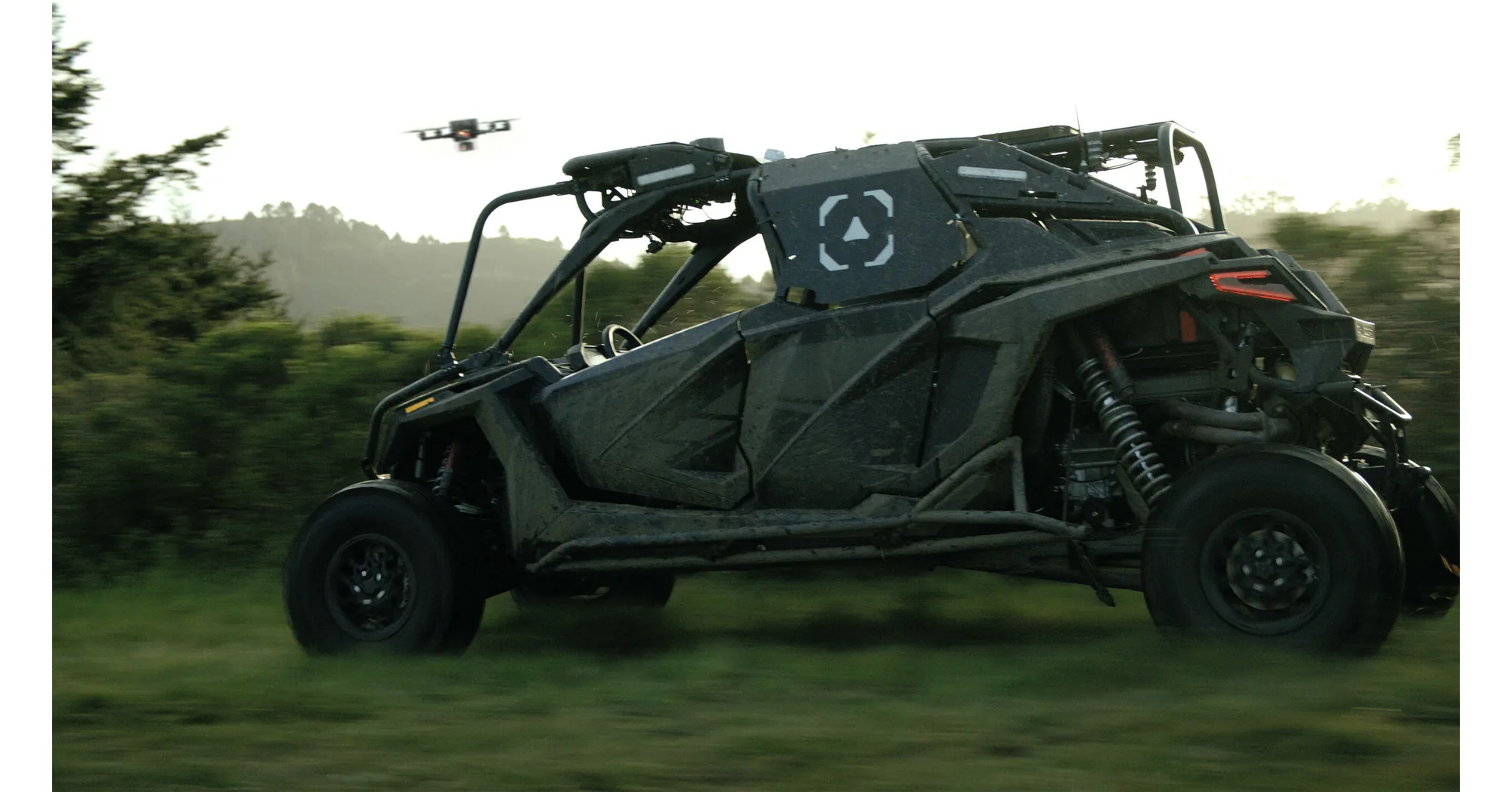 A Scout AI autonomous vehicle driving off-road with a drone overhead